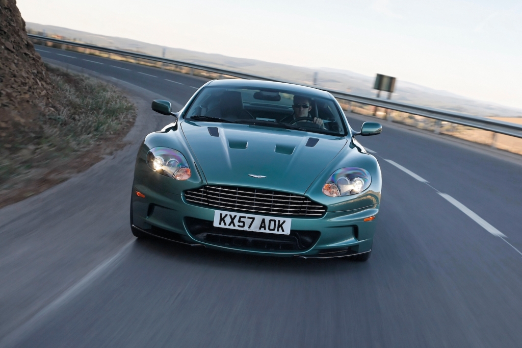 Esta es una foto publicitaria de un Aston Martin DBS V12 gran turismo verde circulando por la autopista. Este es un coche que pertenece tanto a James Bond como a Jay Leno.